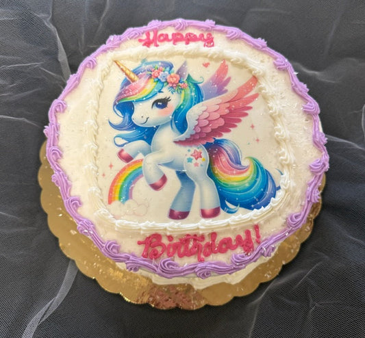 A round two-layer cake with white frosting, purple piped border, "Happy Birthday" in pink cursive frosting, and a printed edible image of a rainbow unicorn with wings and flowers, on a gold board.