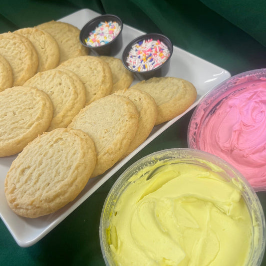 A cookie decorating kit on a green cloth background, including a tray of unfrosted round sugar cookies, containers of pink and yellow frosting, and two small bowls of rainbow sprinkles and jimmies.