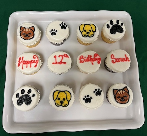 A platter of dog-themed cupcakes with white frosting, featuring illustrated dog faces, paw prints, and a "Happy 12th Birthday Sarah" message spelled across four cupcakes in red frosting.