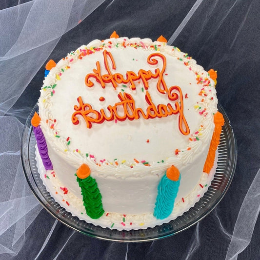 A two-layer, eight-inch round birthday cake. The top reads 'Happy Birthday' in orange and red icing. Decorated with white icing, icing-sculpted candles around the perimeter, and colorful sprinkles.