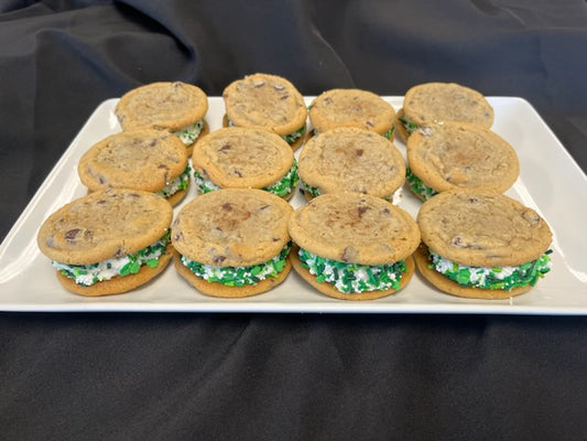 Chocolate chip cookie whoopie pies with fluffy frosting filling rolled in mini green sprinkles, sandwiched between two chocolate chip cookies, arranged on a white rectangular plate against a black background.