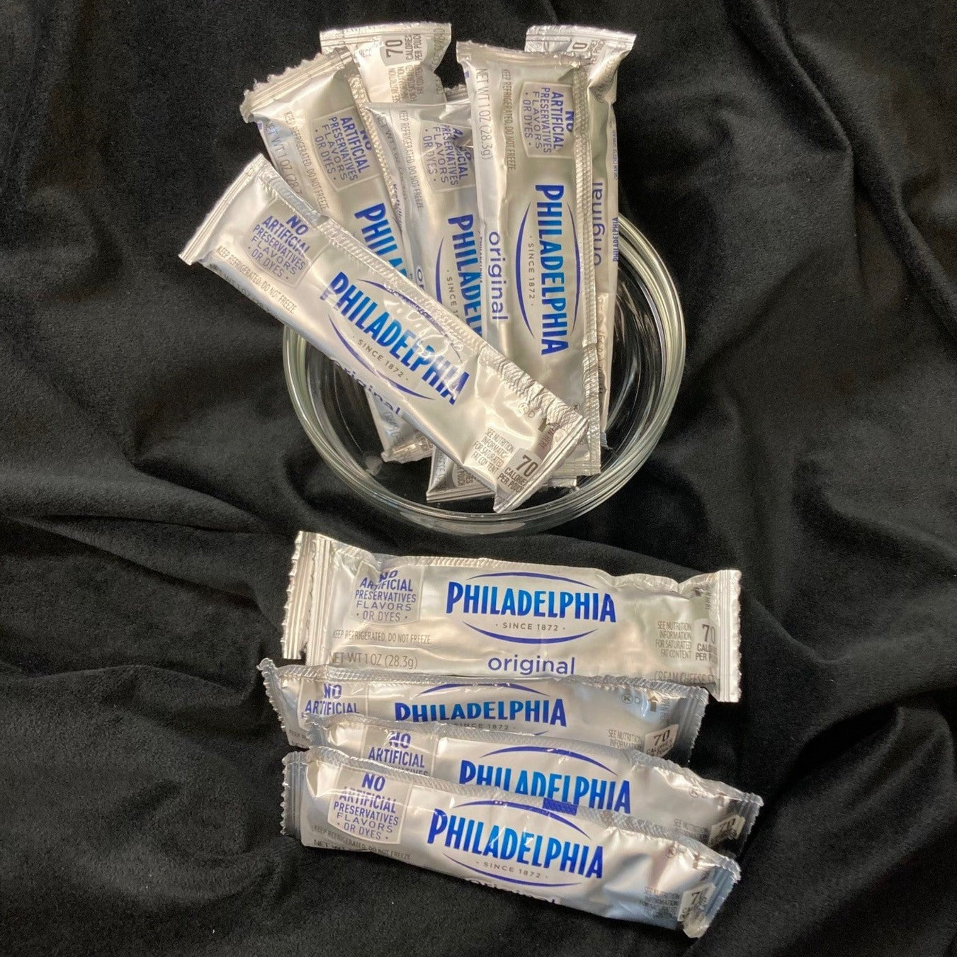 Individual Philadelphia original cream cheese packets in silver packaging, with several arranged in a glass bowl and additional packets scattered on a black fabric surface.