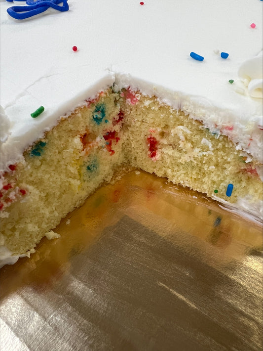 A close-up of a sliced confetti half sheet cake showing a light yellow funfetti interior with colorful sprinkles baked throughout, topped with white frosting.