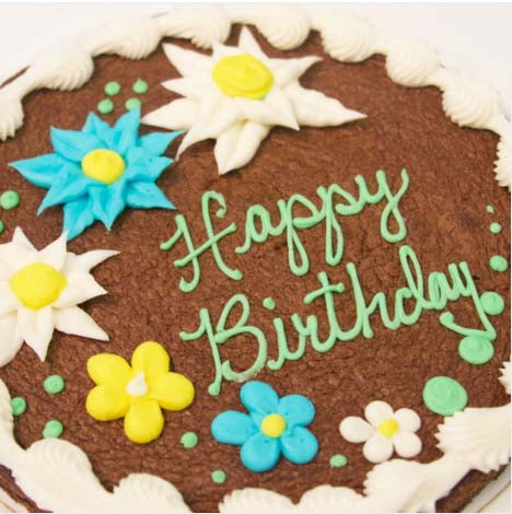 A close-up of a round nine-inch decorated brownie reading 'Happy Birthday' in pastel green cursive icing, with a variety of decorated flowers in blue, yellow, and white, and green polka dots surrounding the flowers. A white shell border runs along the outer edge. The brownie sits on a white cake board against a white background.