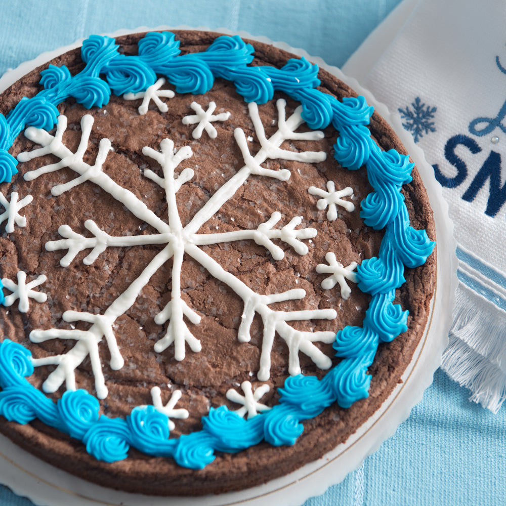 A round nine-inch decorated brownie featuring a large decorated snowflake in the center, surrounded by smaller snowflakes in white icing. A thick blue border runs along the outer edge, with white iridescent sprinkles throughout. The brownie sits on a white cake plate, against a blue background and a winter themed cloth.