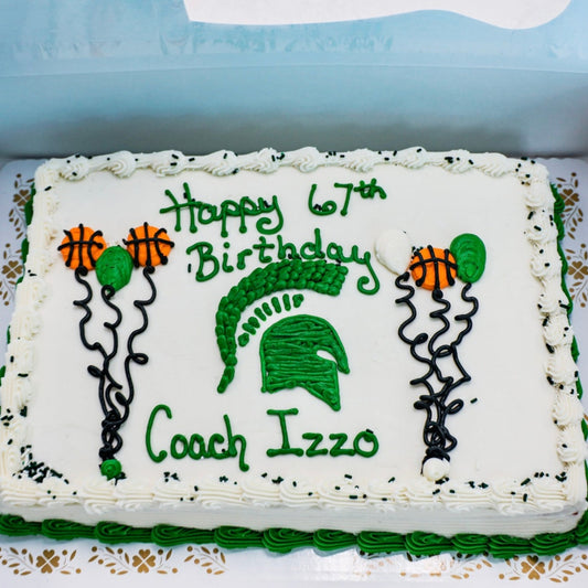 A half-sheet cake with white frosting reading "Happy 67th Birthday Coach Izzo" in green icing, decorated with a Spartan helmet logo, basketball balloon designs, and black sprinkles sprinkled around the edge of the cake.