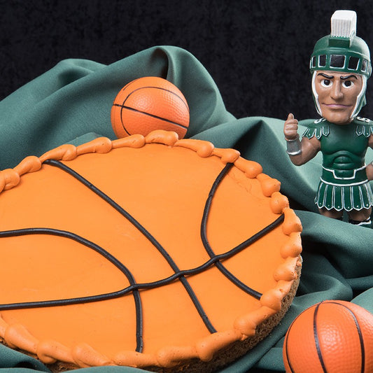A round decorated cookie designed to resemble a basketball, covered in orange icing with black curved lines representing the seams. An orange rope-style icing border runs along the outer edge. The cookie is displayed on dark green fabric alongside two small rubber basketballs and a MSU Sparty bobblehead figurine against a dark background.
