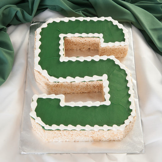 Letter S shaped cake with smooth green frosting on top, white piped frosting outlining the shape, and visible textured cake sides, displayed on a silver board with green fabric in the background.