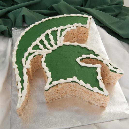A Rice Krispie treat cut and shaped into the Michigan State University Spartan helmet logo, decorated with green frosting and white piping, displayed on a silver board.