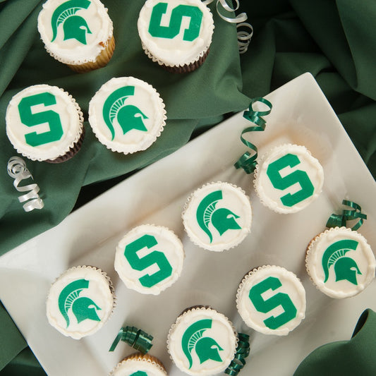 An assortment of cupcakes topped with white frosting printed with Michigan State University logos including the block S and Spartan helmet in green, arranged on a white plate and green linen with curled ribbon accents.