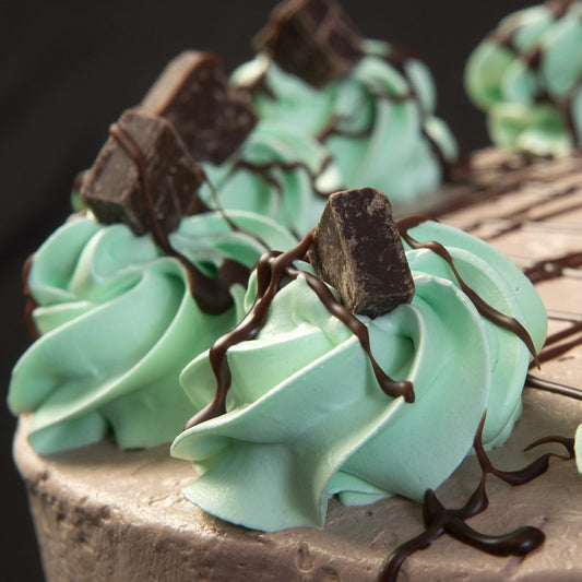 A close-up of mint green whipped topping rosettes topped with chocolate chunks and chocolate drizzle along the edge of a chocolate frosted cake.