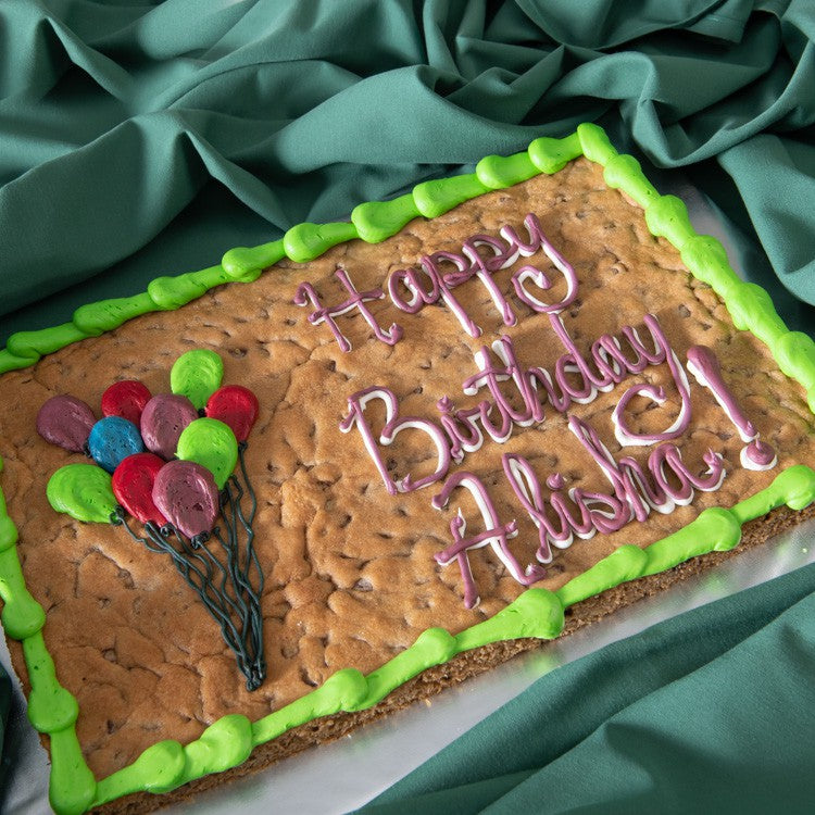 A half sheet chocolate chip cookie with a green border, decorated with colorful balloon clusters and the text "Happy Birthday Alisha!" in pink script, displayed on green linen.
