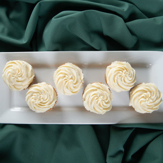 A top view of six vanilla vegan cupcakes topped with a fluffy vanilla frosting, arranged together on a white plate, with a green background.