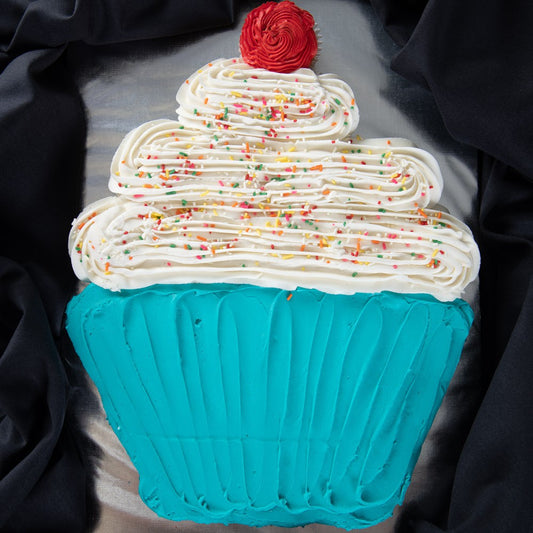 A cupcake-shaped cake decorated with teal frosting on the base, white swirled frosting with rainbow sprinkles on top, and a red rosette, displayed against black fabric.