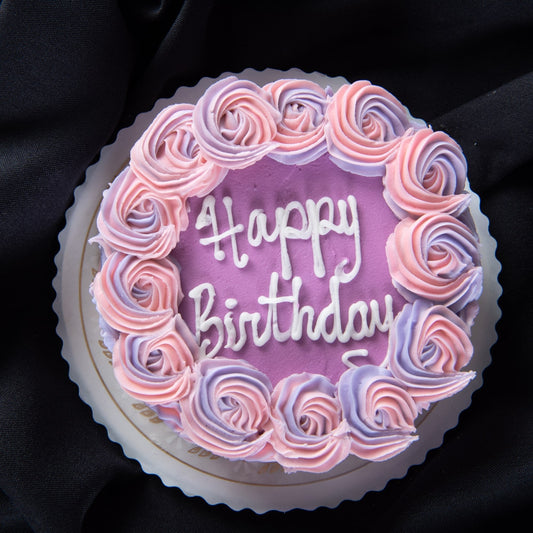 A round cake with a pink frosted top decorated with pink and purple rosettes around the border and "Happy Birthday" written in white script, displayed on a cake board against black fabric.