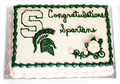 A sheet cake decorated with white and green icing featuring "Congratulations Spartans" text, a Spartan S logo, a MSU Spartan brand helmet, and white flowers with green leaves.