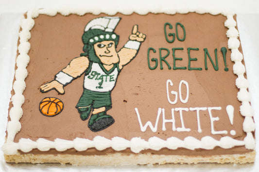 A rectangular Rice Krispie treat with chocolate frosting and white border, decorated with the Michigan State University Sparty mascot dribbling a basketball and the text "Go Green! Go White!" in green and white.