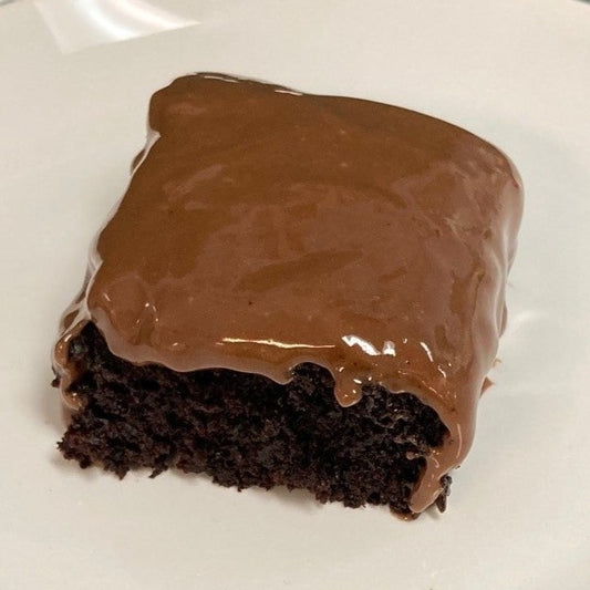 A single square slice of vegan chocolate cake with a dense, moist dark chocolate crumb topped with glossy chocolate topping on a white plate.