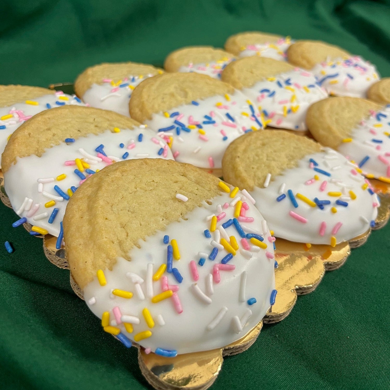 A stack of dipped sugar cookies half-coated in white chocolate and topped with pink, blue, yellow, and white sprinkles, displayed on a gold board against a green cloth background.