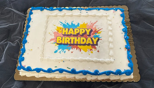 A rectangular half sheet cake with white frosting, blue piped border detailing, rainbow sprinkles, and a printed edible image reading "Happy Birthday" in bold yellow and red text with colorful paint splatter design, on a gold board.