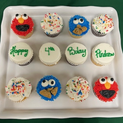 A platter of Sesame Street-themed cupcakes featuring Elmo faces with red frosting, Cookie Monster faces with blue frosting and crumbled cookie pieces, white frosting topped with rainbow sprinkles, and a "Happy 4th Bday Andrew" message spelled across four cupcakes in green frosting.