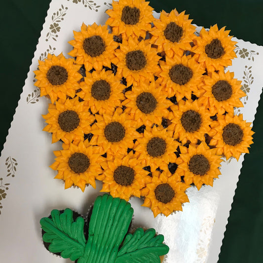A pull-apart cupcake cake arranged in the shape of a sunflower bouquet. Approximately 20 cupcakes are decorated with orange-yellow petal frosting and dark brown textured centers to mimic sunflowers. Green frosted stems and leaves are visible at the bottom of the arrangement, set on a white board.